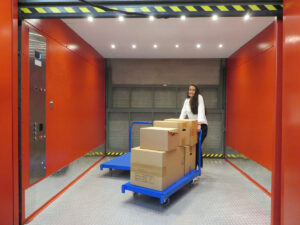 A woman in a centron storage building stands in a red elevator pushing a cart filled with boxes.