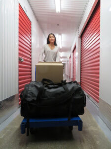 A woman is pushing a cart that holds moving boxes and personal belongings, while walking through the hallway of Centron Storage.